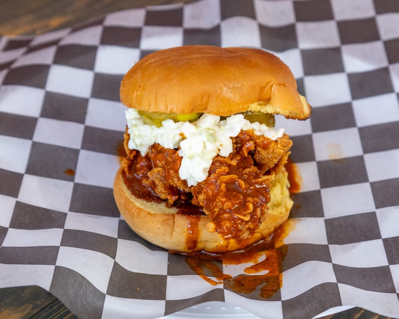 Saucy fried chicken sandwich with coleslaw and pickles on a bun, on checkered paper.
