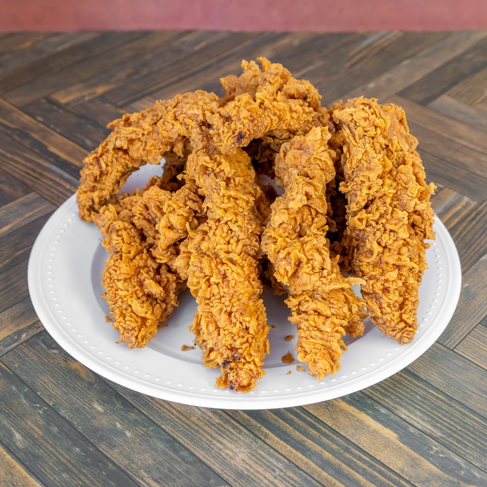Pile of crispy, golden-brown fried chicken tenders on a white plate set on a wooden table.