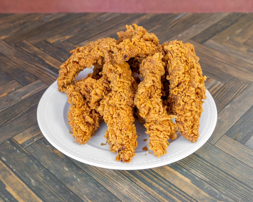 Pile of crispy, golden-brown fried chicken tenders on a white plate set on a wooden table.