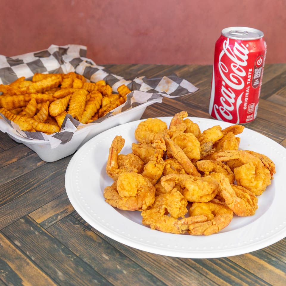 Plate of golden fried shrimp, crinkle-cut fries in a basket, and a Coca-Cola can on a wooden table.