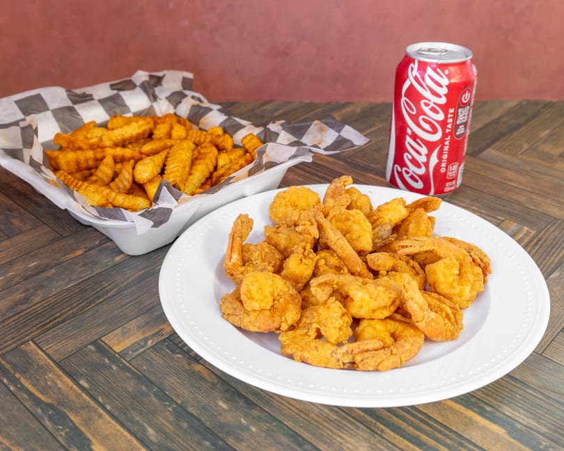 Plate of golden fried shrimp, crinkle-cut fries in a basket, and a Coca-Cola can on a wooden table.