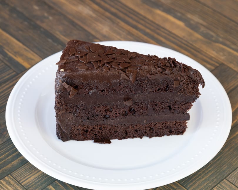 Slice of rich chocolate cake with chocolate frosting and shavings on a white plate.