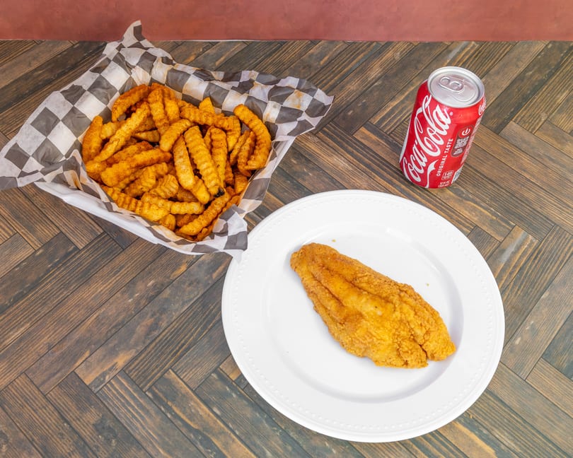 Fried fish fillet, seasoned crinkle-cut fries in a basket, and a Coca-Cola can on a wooden table.