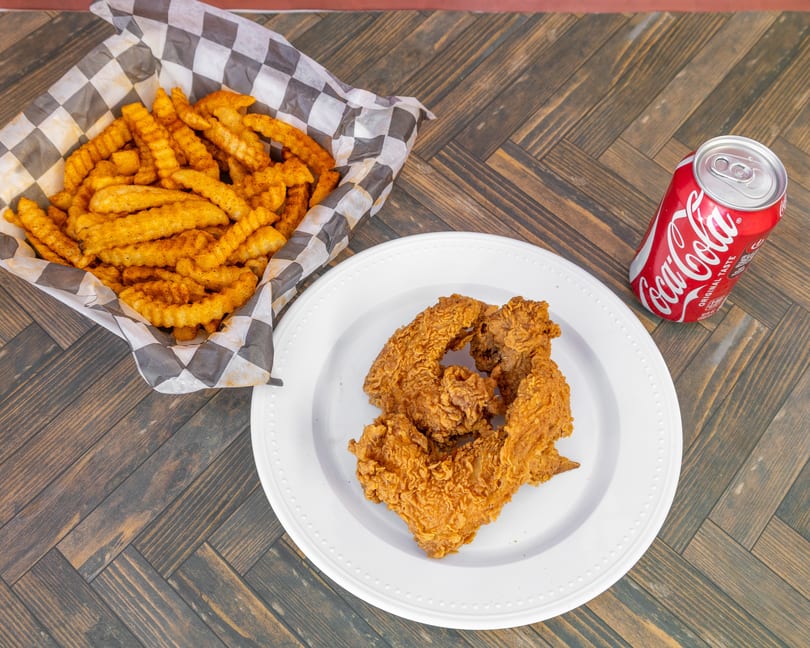 Fried chicken on a white plate, crinkle-cut fries in a basket, and a Coca-Cola can on a wooden table.