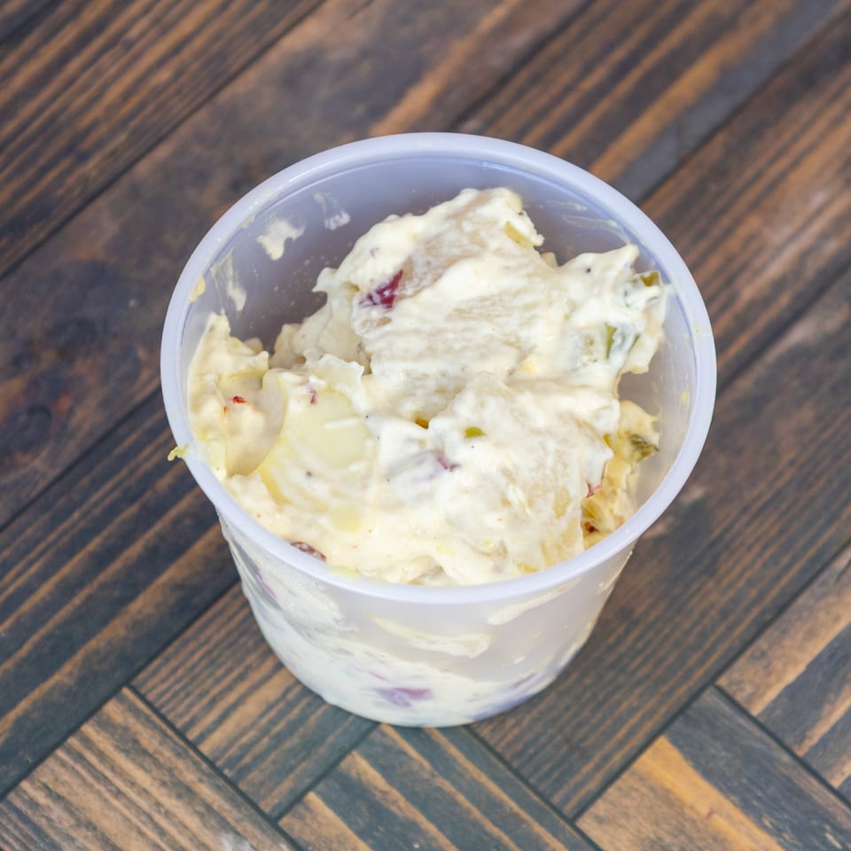 Creamy potato salad with red and green bits in a plastic cup on a wooden table.