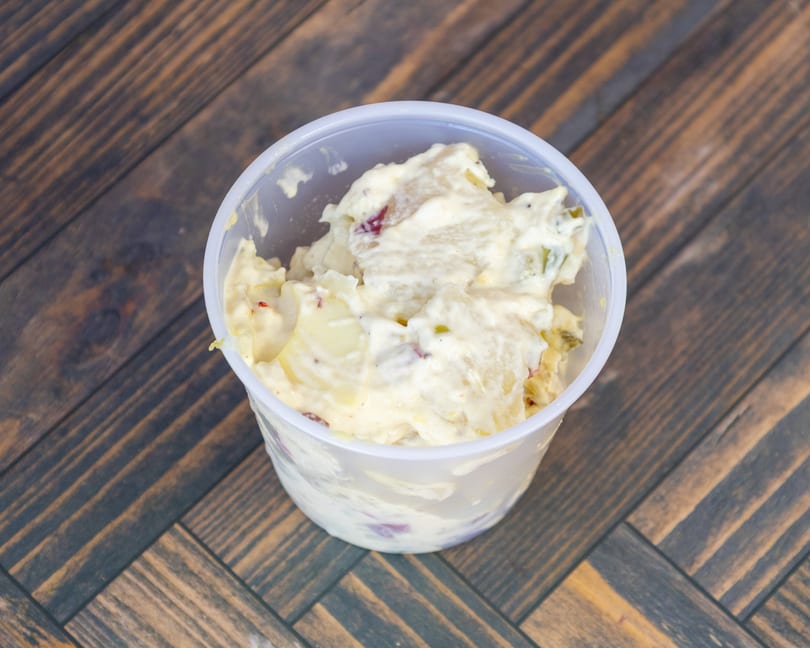 Creamy potato salad with red and green bits in a plastic cup on a wooden table.