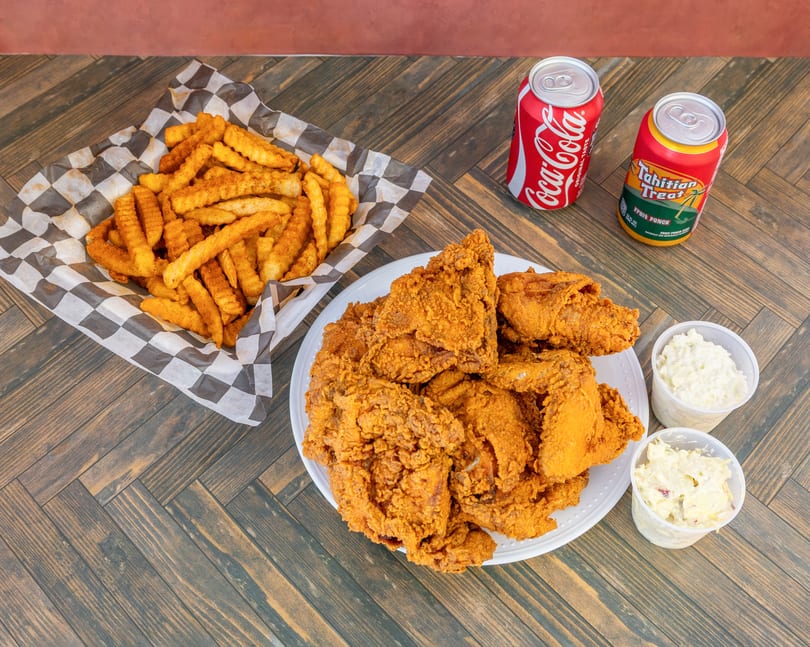 Fried chicken, waffle fries, coleslaw, potato salad, Coca-Cola, and Tahitian Treat on a wooden table.