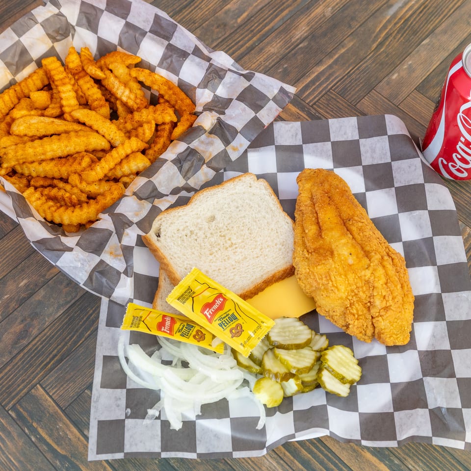 Fried fish fillet, crinkle fries, bread, pickles, onions, mustard, cheese, and a Coca-Cola on wood.