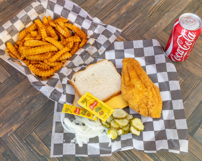 Fried fish fillet, crinkle fries, bread, pickles, onions, mustard, cheese, and a Coca-Cola on wood.