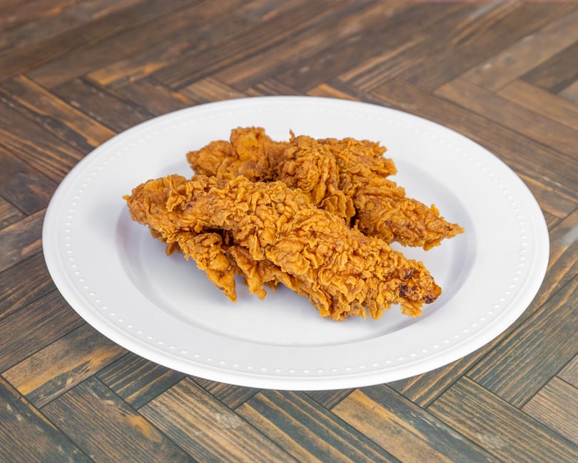 Crispy golden brown fried chicken tenders on a white plate set on a wooden table.
