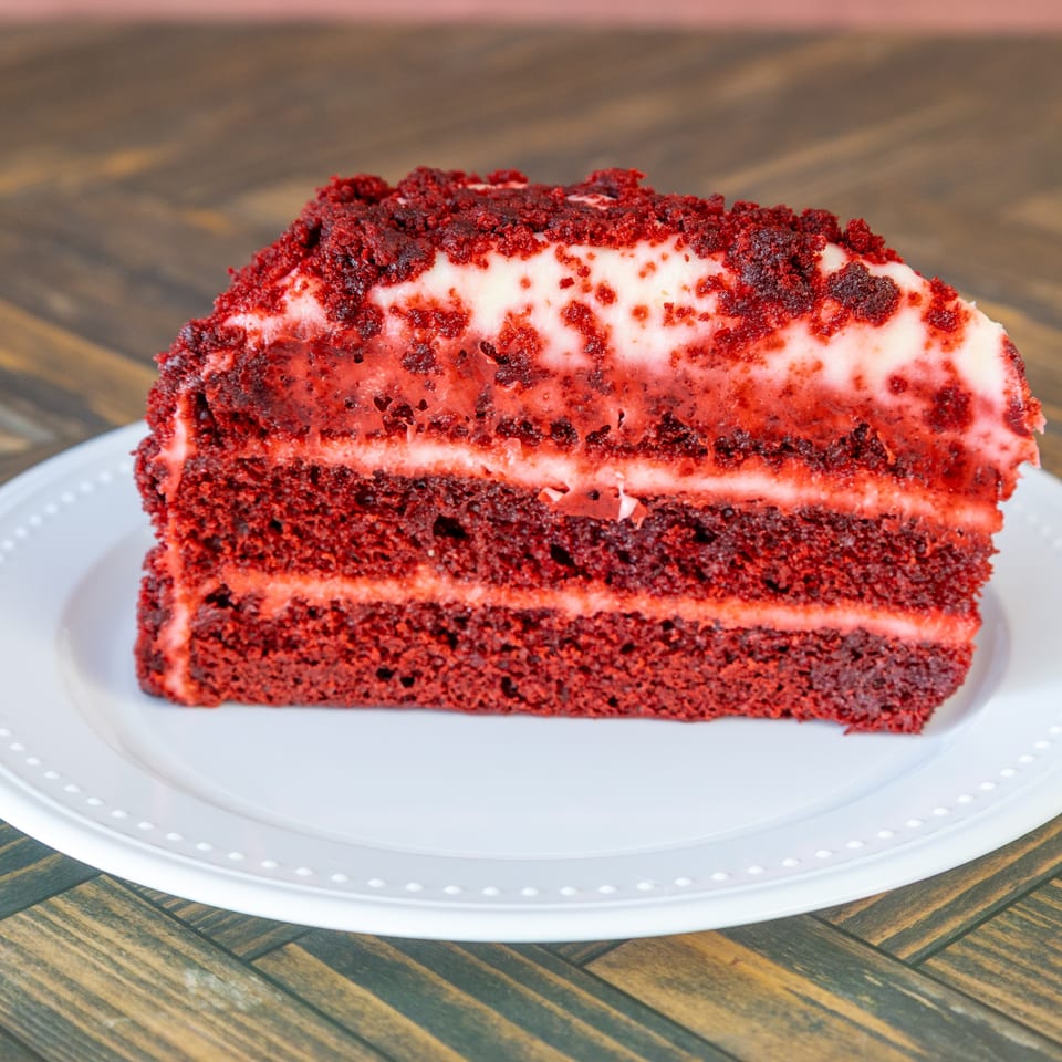 A slice of red velvet cake with white frosting layers and crumb topping on a white plate.