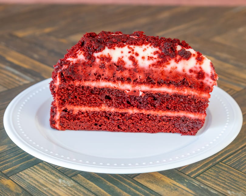 A slice of red velvet cake with white frosting layers and crumb topping on a white plate.