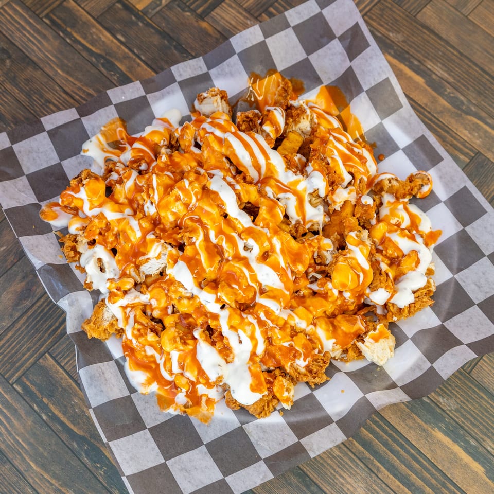 Crispy chicken and fries drizzled with buffalo and ranch sauces on checkered paper.