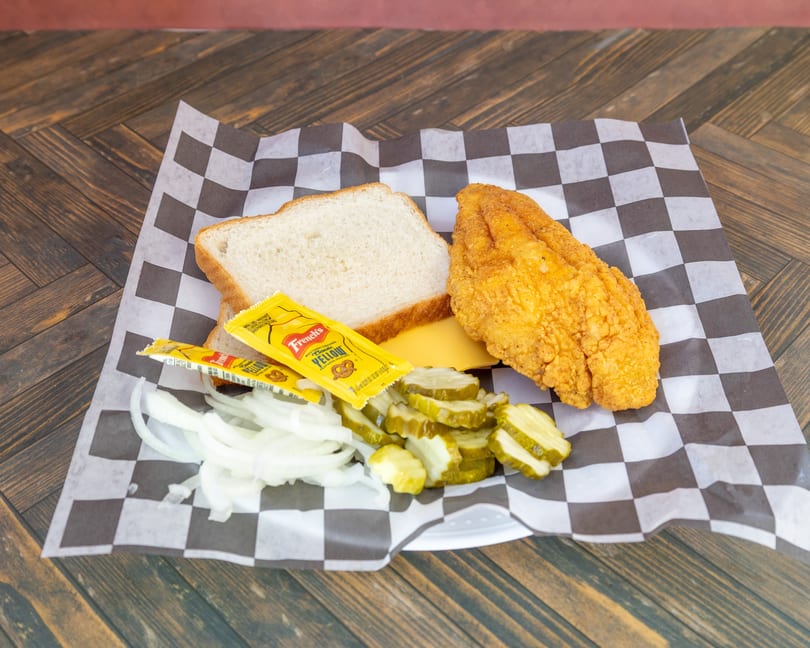 Deconstructed fried fish sandwich with bread, cheese, onions, pickles, and mustard on checkered paper.