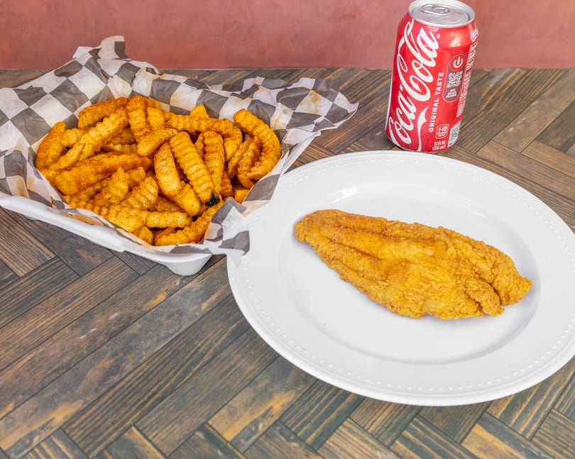 Fried fish fillet, seasoned crinkle-cut fries in a basket, and a Coca-Cola can on a wooden table.