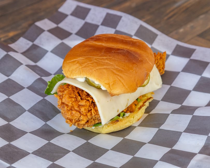 Crispy fried chicken sandwich with cheese, lettuce, pickles, and sauce on a bun, on checkered paper.
